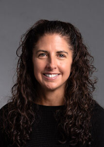 An adult with long dark curly hair and peach colored skin smiles at the camera